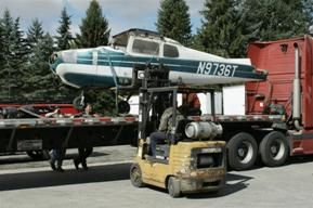 A forklift lifts a small airplane off a flatbed truck in a loading area surrounded by trees.