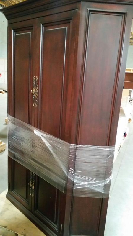 Wooden cabinet with a rich brown finish, featuring two doors and decorative handles, wrapped in plastic for protection.