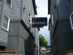 Narrow alleyway between two gray buildings with a sign hanging overhead and greenery visible in the background.