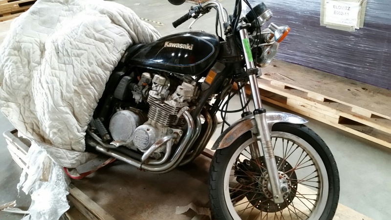 A black Kawasaki motorcycle is partially covered with a quilt, resting on wooden pallets in a warehouse.