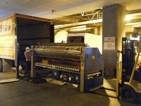 A large printing press being unloaded from a truck in an industrial setting, with a forklift nearby.