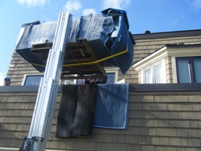 A lift is positioned near a house, with a covered object being hoisted above the roof, surrounded by blue sky and windows.