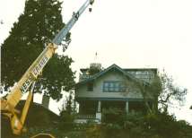 A crane is lifting materials towards a two-story house surrounded by trees.