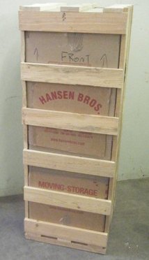 A wooden crate with slatted sides, labeled "Hansen Bros. Moving Storage," standing upright in a storage area.