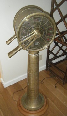 A vintage brass pedestal device with dials and two telescopic lenses, set on a wooden base in a room.